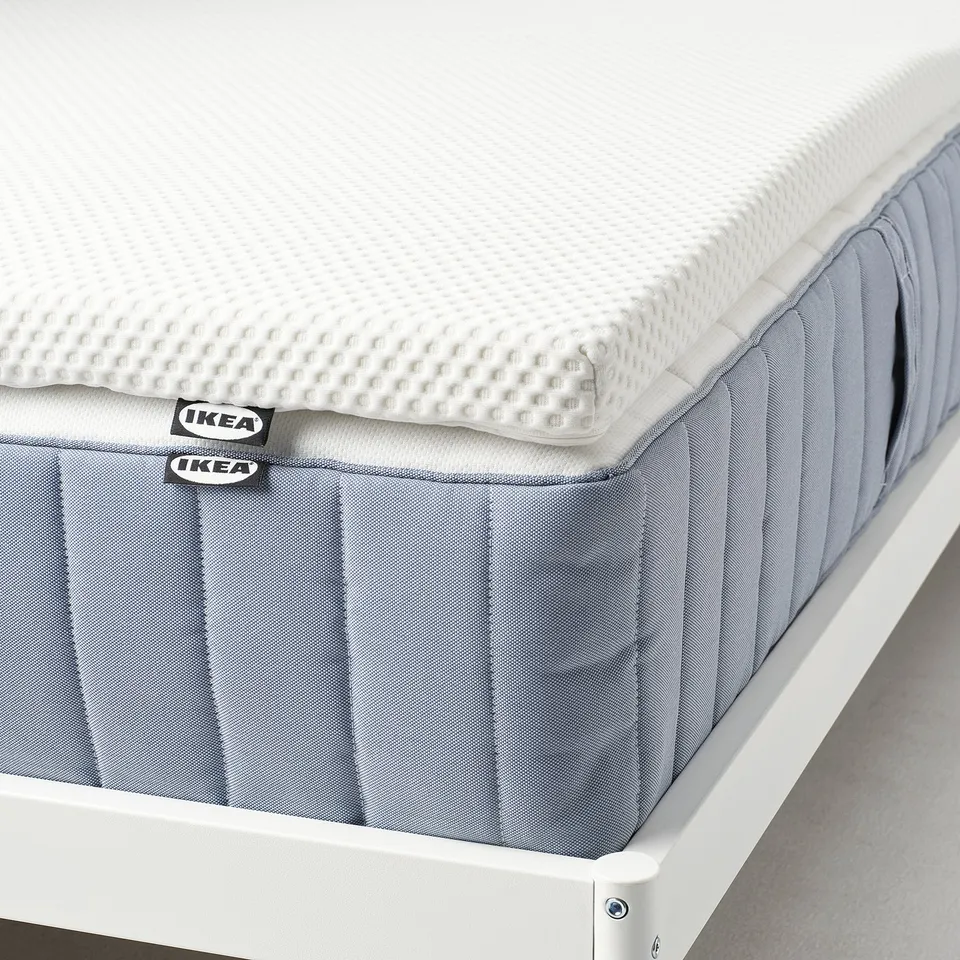 Close-up of a roll-packed IKEA mattress pad featuring pressure-relieving memory foam, displayed on a white bed frame.