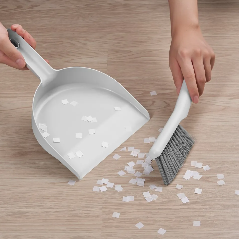 Person uses PEPPRIG dustpan and brush to sweep up small squares on wooden floor. Brush is snapped onto dustpan handle.