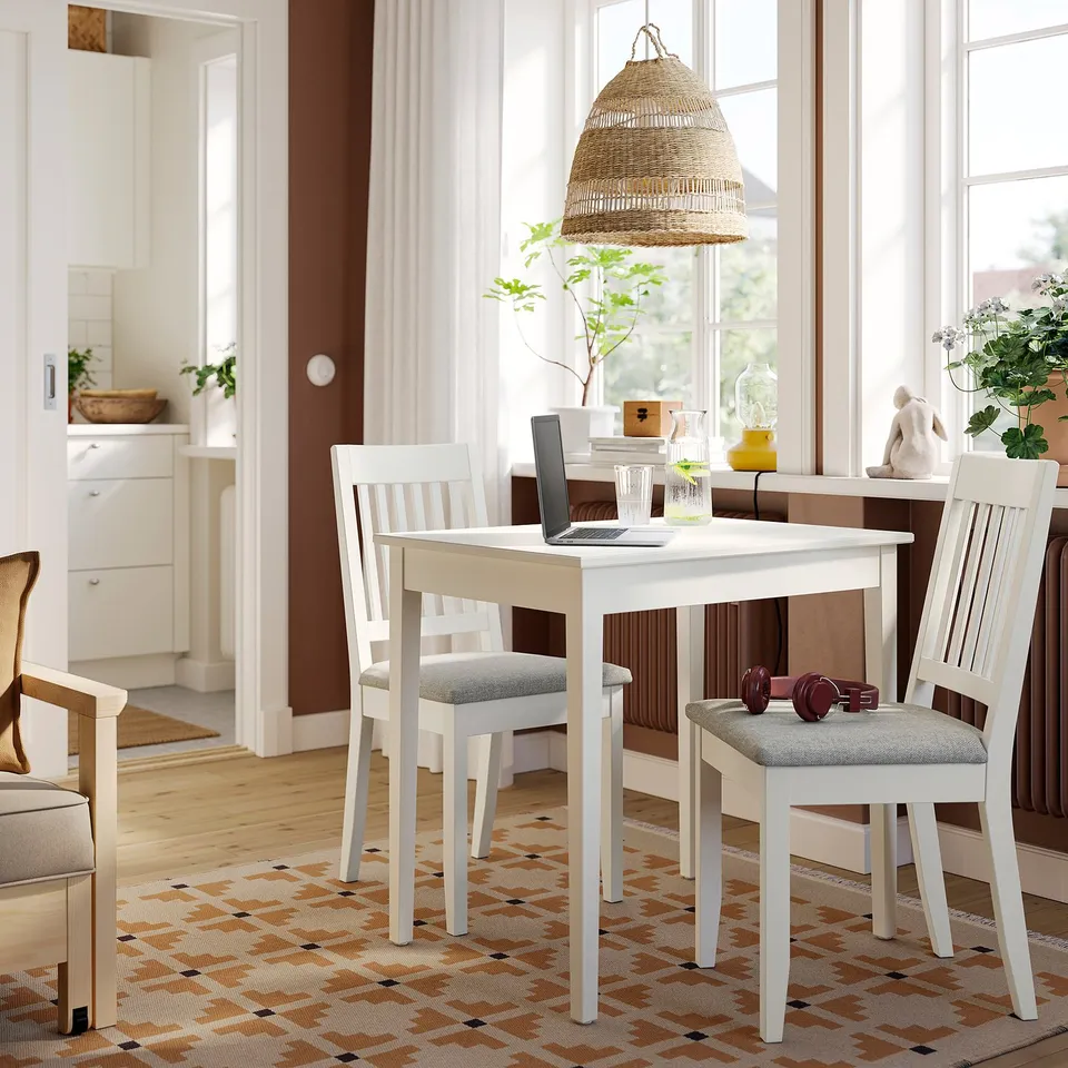 Small white NÄSINGE dining set with cushioned chairs fits neatly under a large window, featuring a light wooden floor and cosy decor.