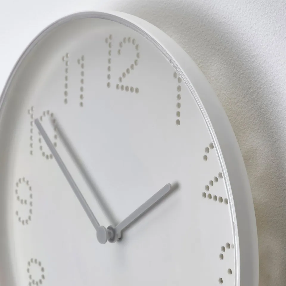 White round wall clock with punched hole numbers, hands pointing at twelve and six. Minimalist design, mounted on a plain wall.