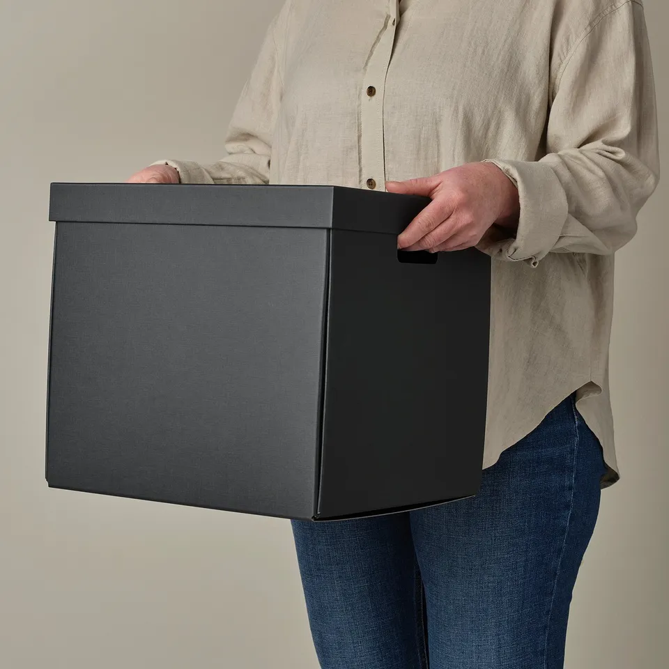 A person holding a square black storage box with cut-out handles, ideal for organising and carrying items.