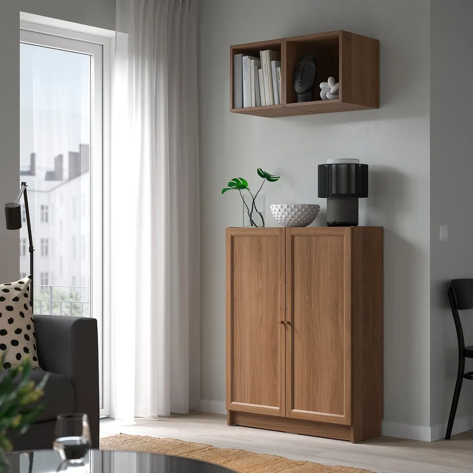 Light wood BILLY bookcase & cabinet set against grey wall; houses books, decor, and plant; size between chair and large window.