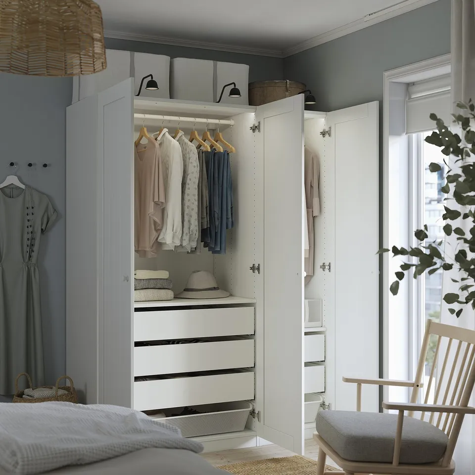 Bedroom with open white wardrobe displaying clothes and drawers, featuring hanging space, shelves, and storage baskets, against blue wall.