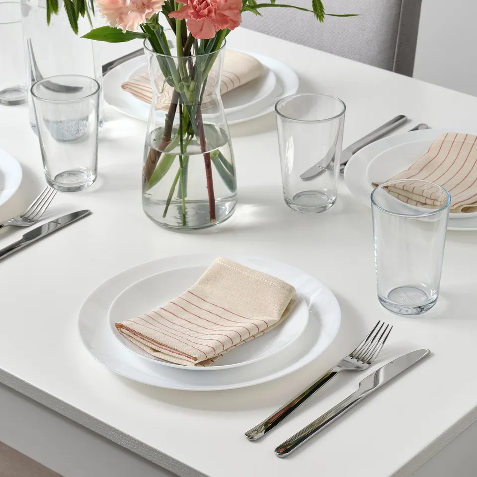 Table set with white plates, clear glasses, silverware, and beige striped napkins. Pink flowers in a vase centrepiece.