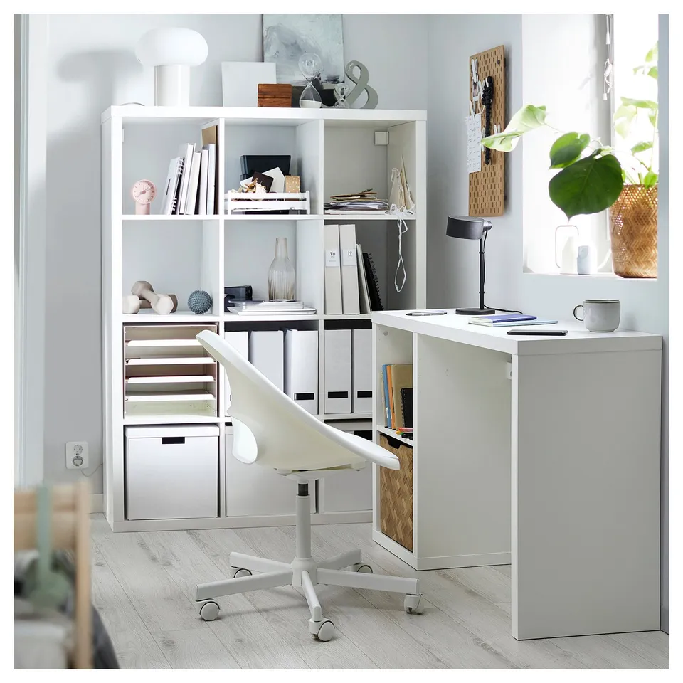 Modern white home office setup with KALLAX bookshelf, desk, and white chair. Shelves hold books, decor, and a lamp. Desk has a computer and resources.