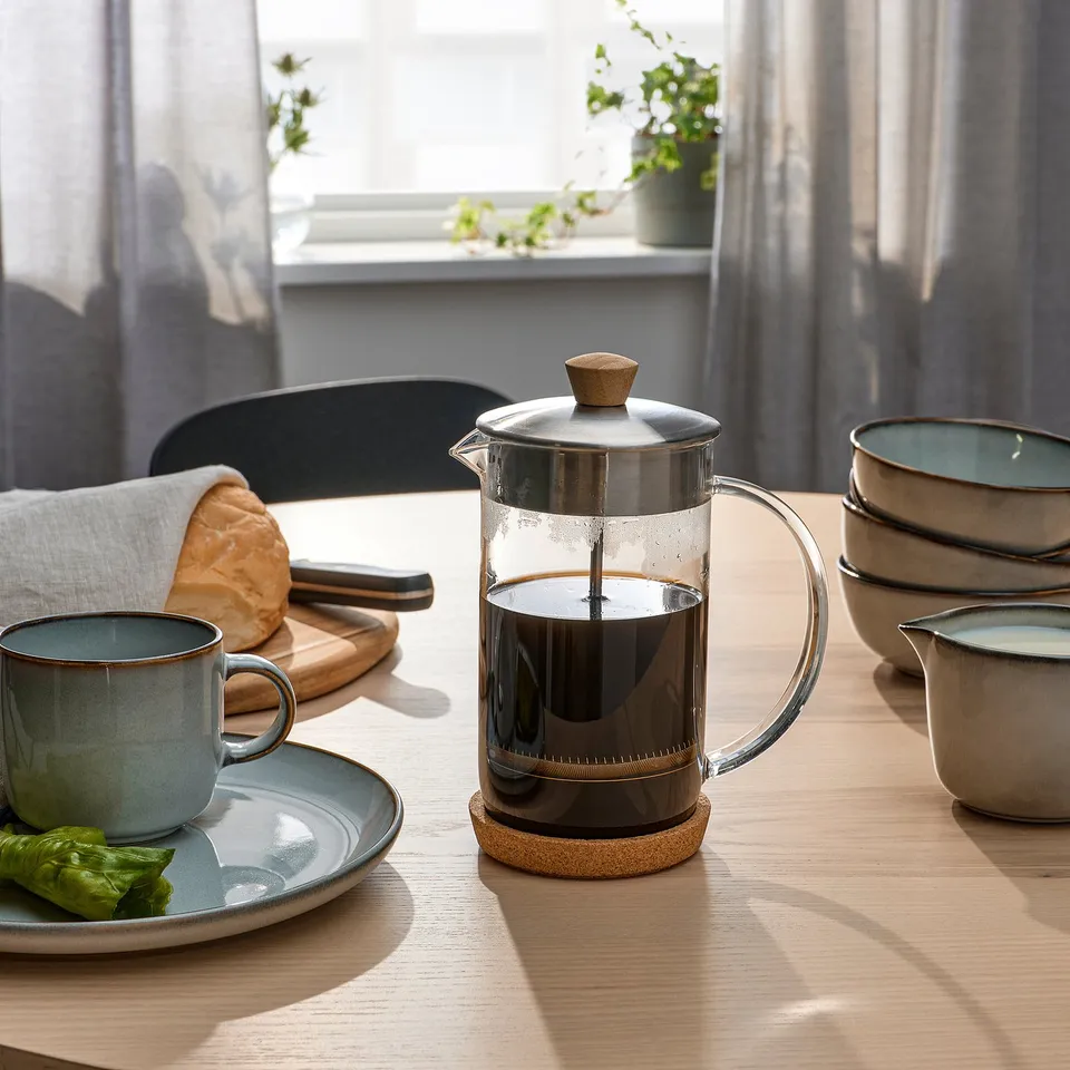A dining table set with a glass coffee maker, mug, plates, and a loaf of bread, ready for breakfast.