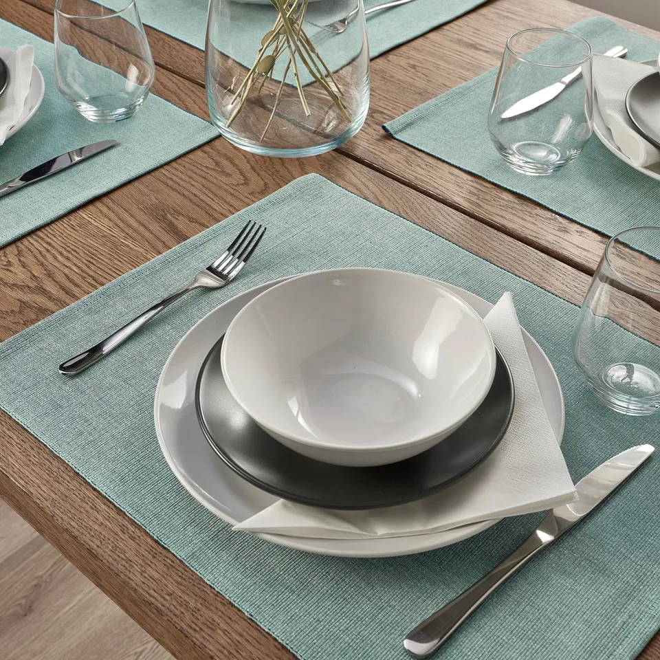 Table setting with green-blue place mats, white plates, bowls, cutlery, and glasses arranged neatly.