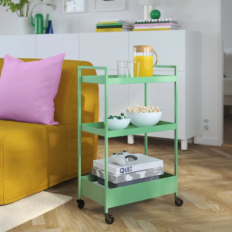 A NISSAFORS green metal trolley near a mustard couch. Small, it holds snacks, games, and drinks, blending vibrantly in a cosy living space.