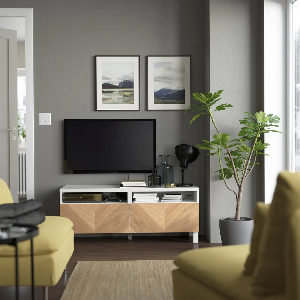 Modern tv room with BESTÅ white & wood tv bench, flat-screen tv, yellow accent chairs, pictures, large plant, lamp, and books.