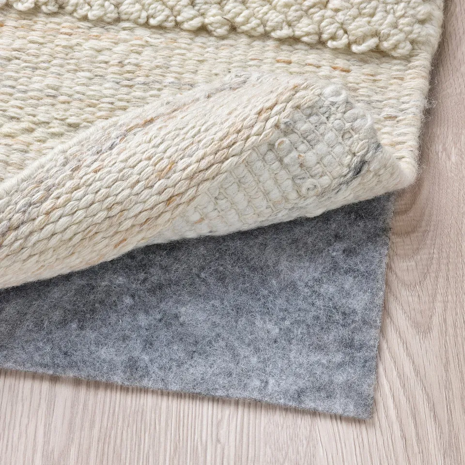 A hand-woven wool rug with a textured pattern. Underneath, theres a grey rug underlay, providing anti-slip stability and comfort.