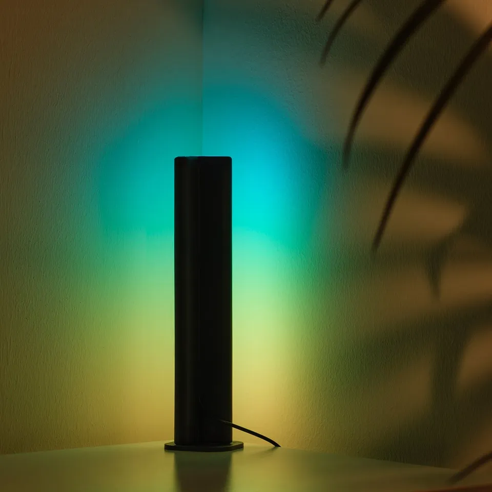 Small black SKAFTSÄRV light emits green and blue hues, sits on white surface.