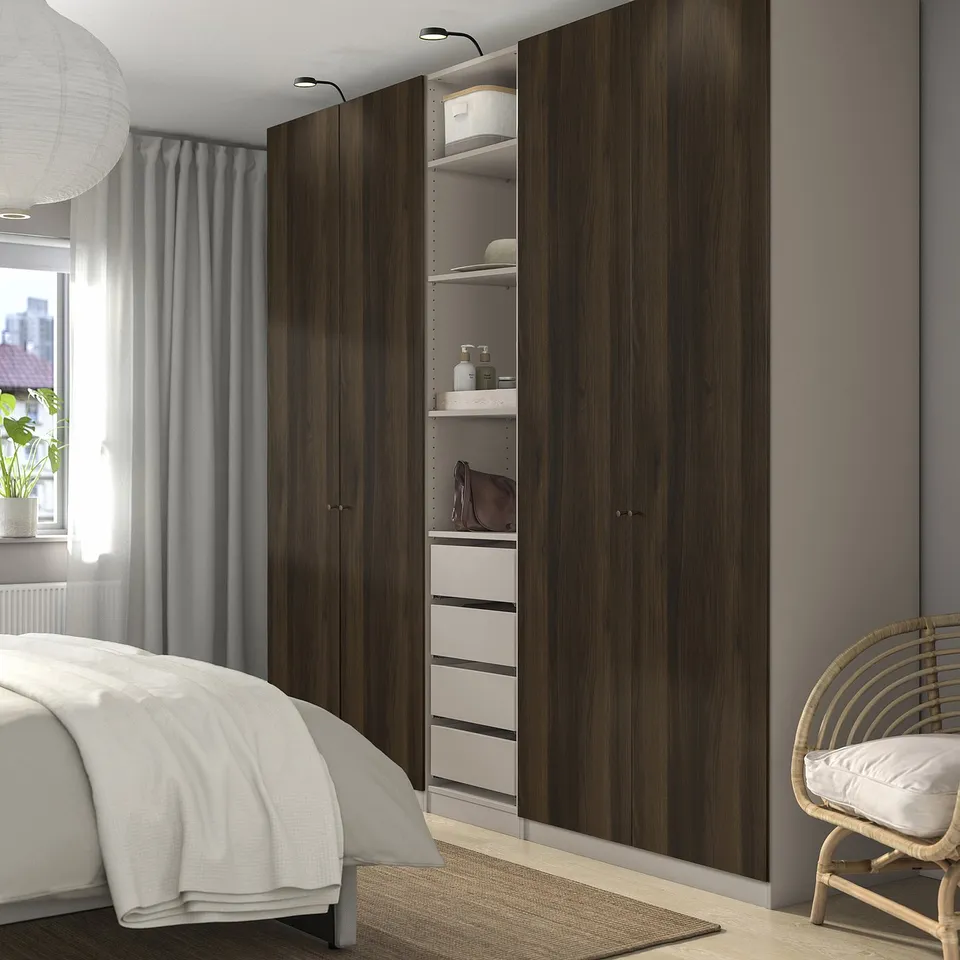 Modern bedroom featuring large PAX wardrobe. Tall, dark wooden closets with open shelves and drawers, positioned beside a white bed and grey curtains.