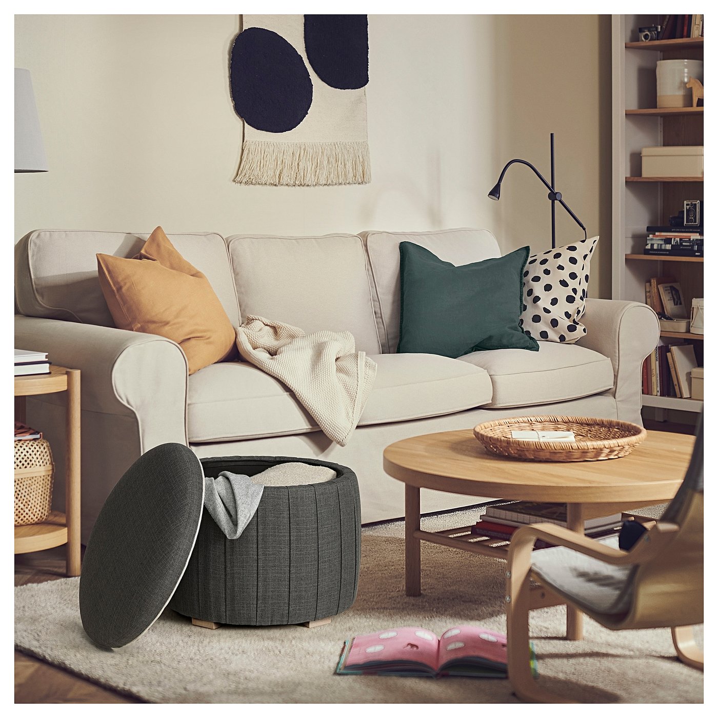 Modern living room with beige sofa, colourful pillows, grey ottoman, wooden coffee table, and bookshelf.