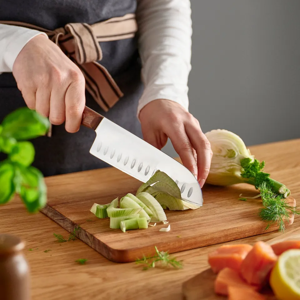 Person slicing fennel with a VARDAGEN knife on a wooden cutting board. Knife has indentations to prevent food from sticking.