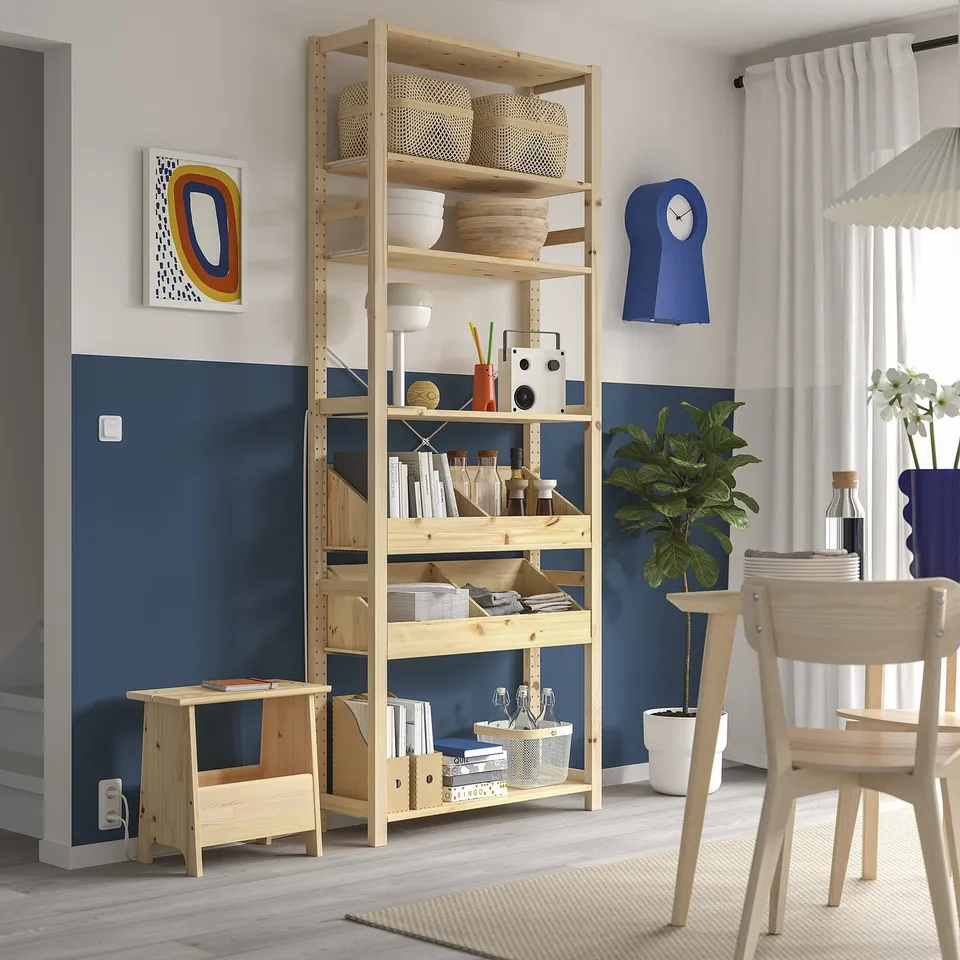 Wooden IVAR shelves hold items, tall and sturdy, light brown. Surrounded by chairs, plants, and decor, fitting room well.