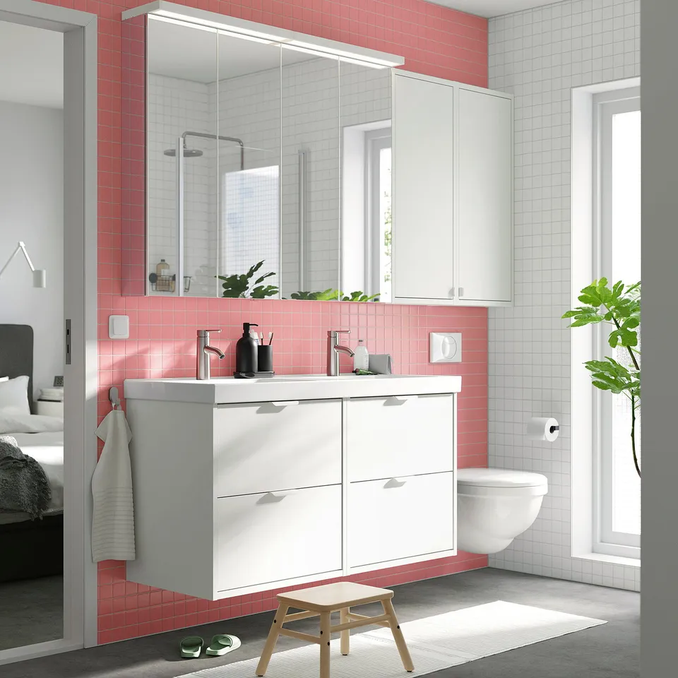 Modern bathroom with HAVBÄCK wall cabinet, pink & white tiles, mirror with lights, plants, and white toilet.
