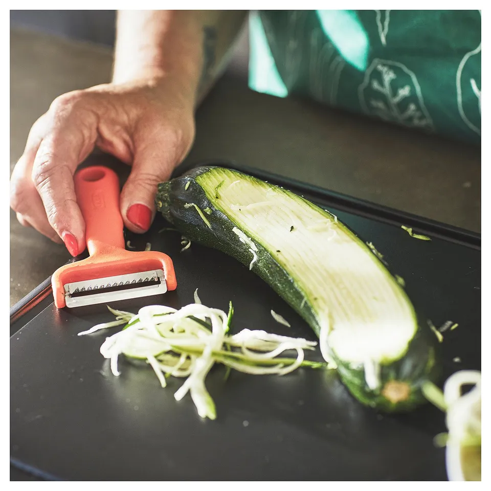 A person uses a red-handled UPPFYLLD vegetable cutter to shred a green cucumber on a black cutting board.