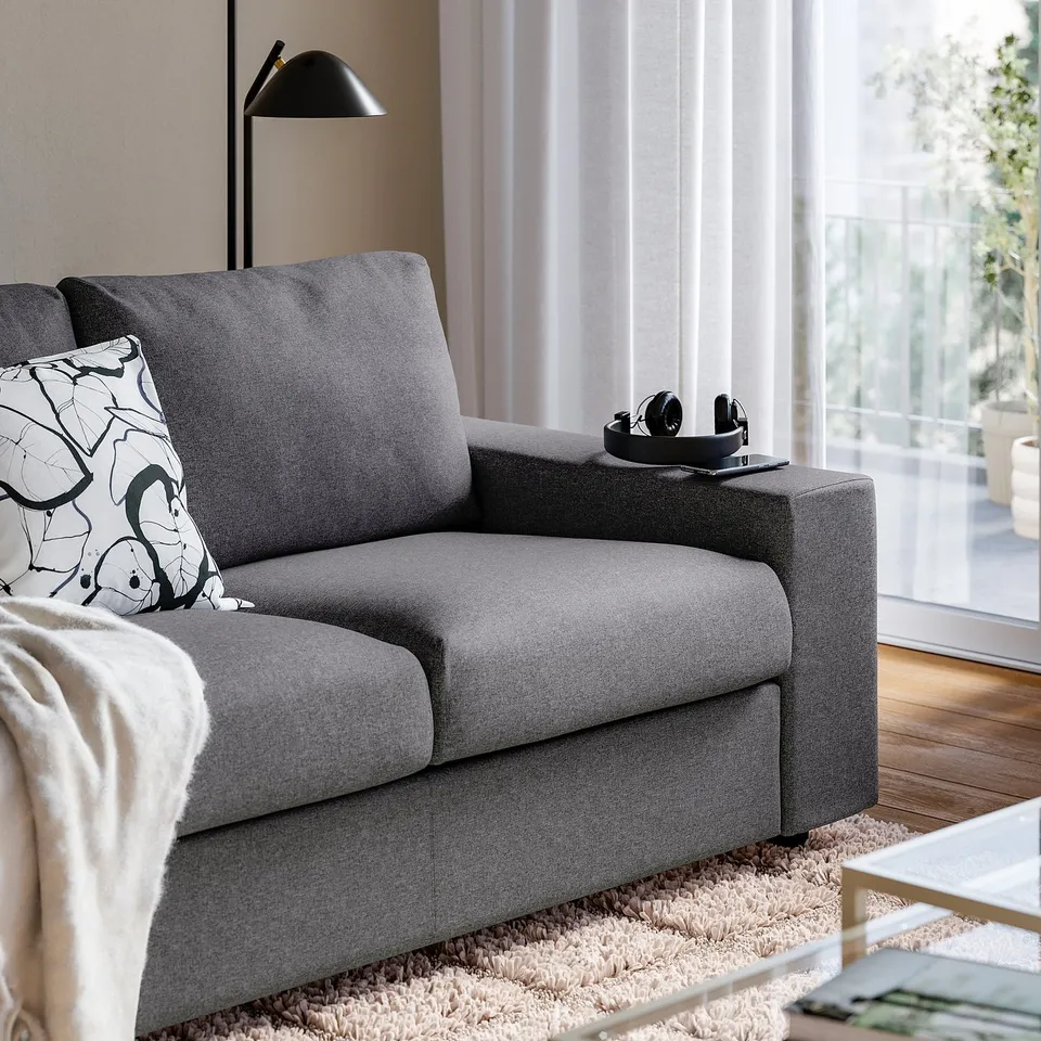 Modern grey sofa with black headphones on arm, soft cushions, by window.