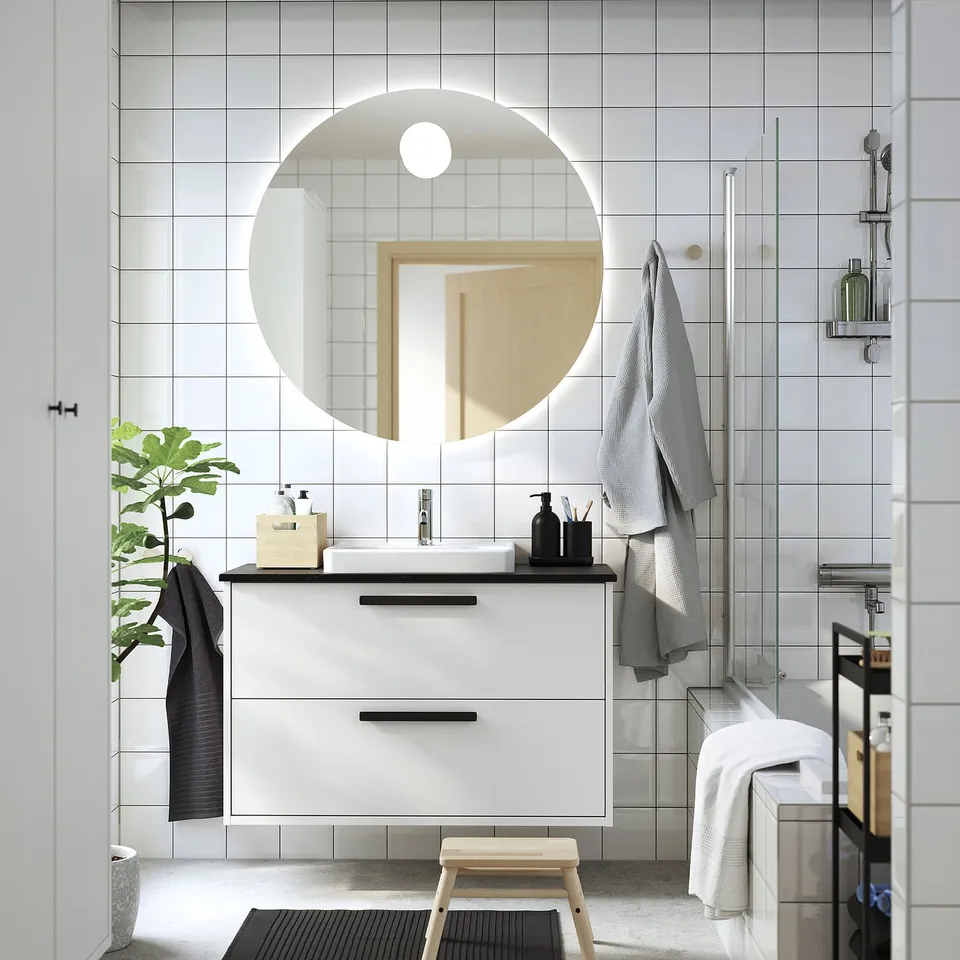 Modern bathroom with round mirror over HAVBÄCK sink, sleek lines, bright white tiles.