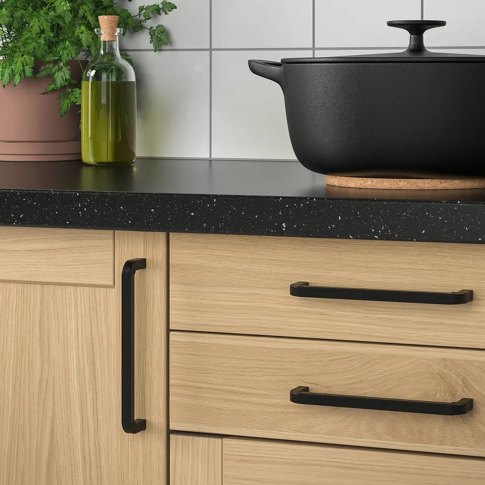 Wooden kitchen cabinet with black iron BORGHAMN handles and knobs, featuring a black countertop with a green bottle and potted herbs.