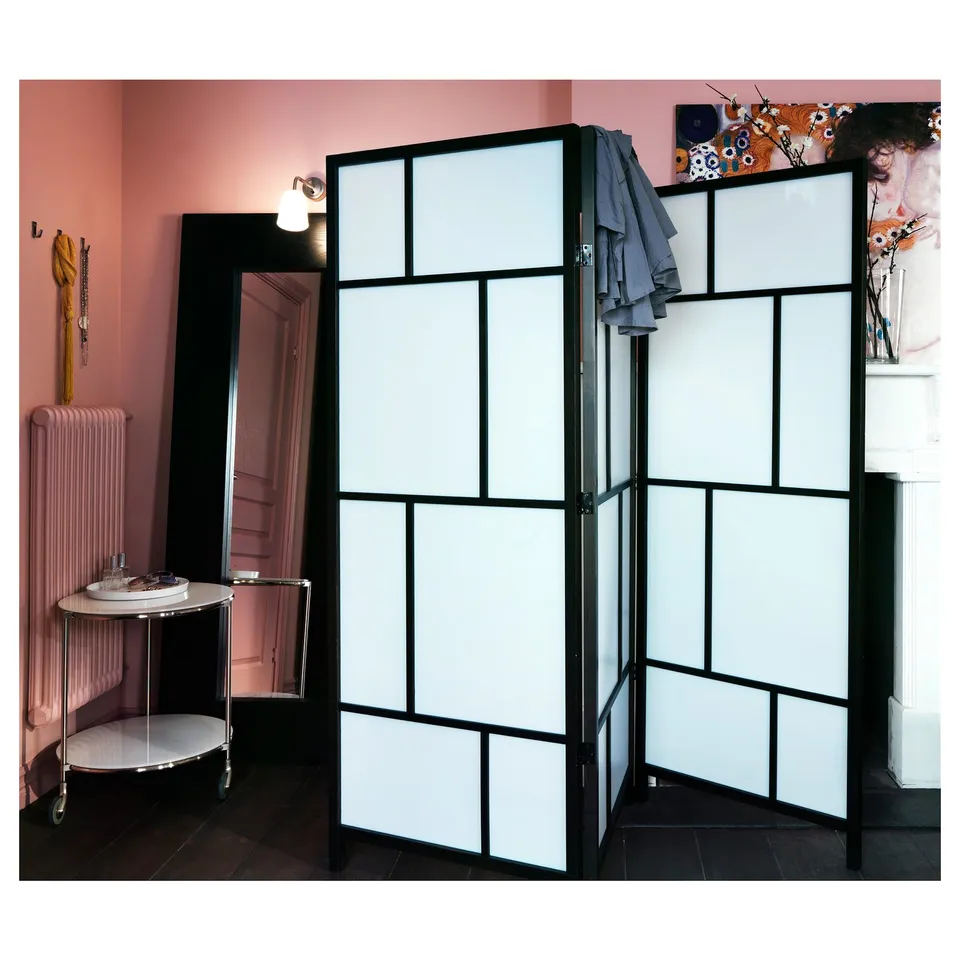 Black room divider with frosted panels creates distinct spaces, positioned in a pink room with a side table, a radiator, and wall decorations.