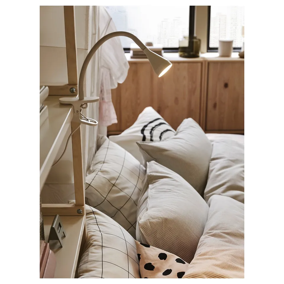 Bedside with cream-colored NÄVLINGE lamp attached to headboard, illuminating a cosy bed with patterned pillows and a wooden dresser in the background.