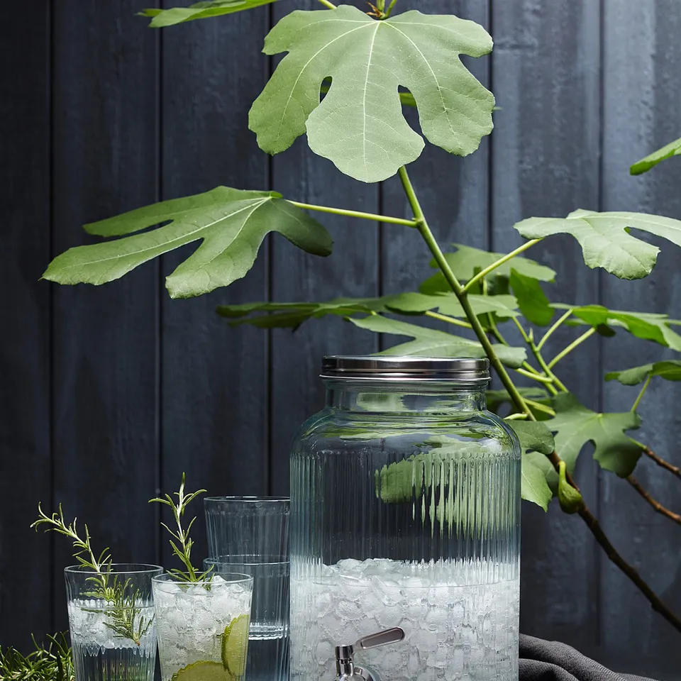 Clear glass VARDAGEN beverage dispenser with stainless steel tap, containing ice cubes. Surrounded by tall glasses with lime slices on dark table.