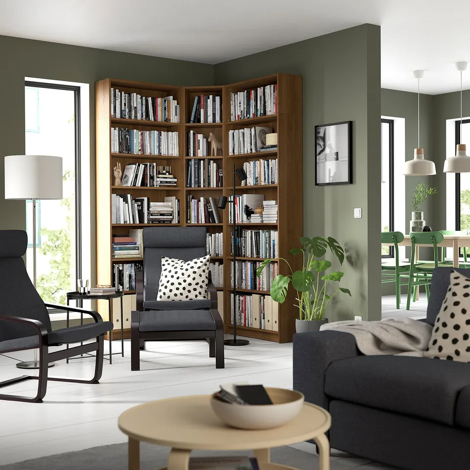 Spacious living room with tall wooden BILLY bookcase filled with books. Dark grey armchairs, grey sofa, and large windows. Green walls and plants.