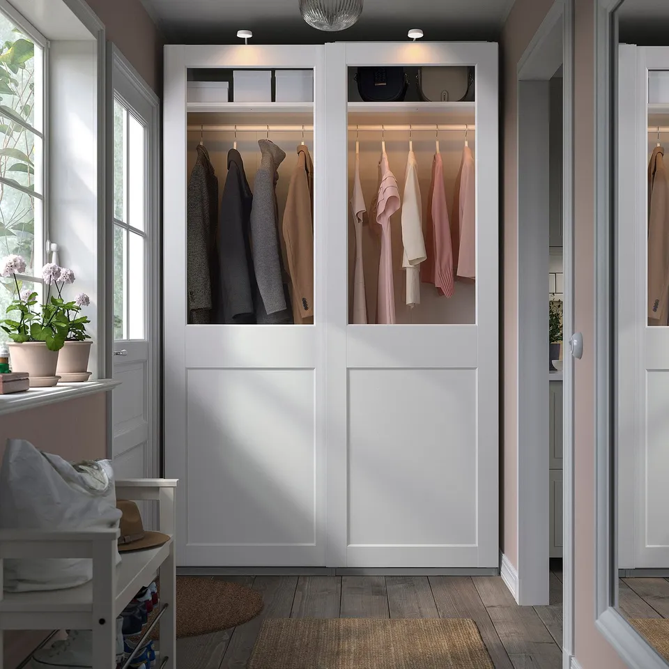 White PAX wardrobe with glass doors displays clothes by windows, next to a small stool with a hat.