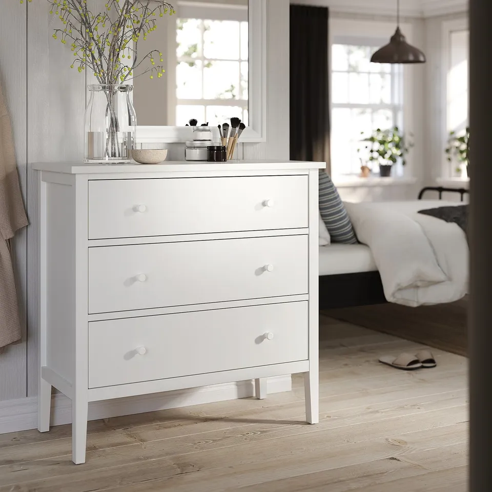 White 3-drawer dresser in bedroom, light wood floor, hanging light, bed with yellow flowers, makeup brushes.