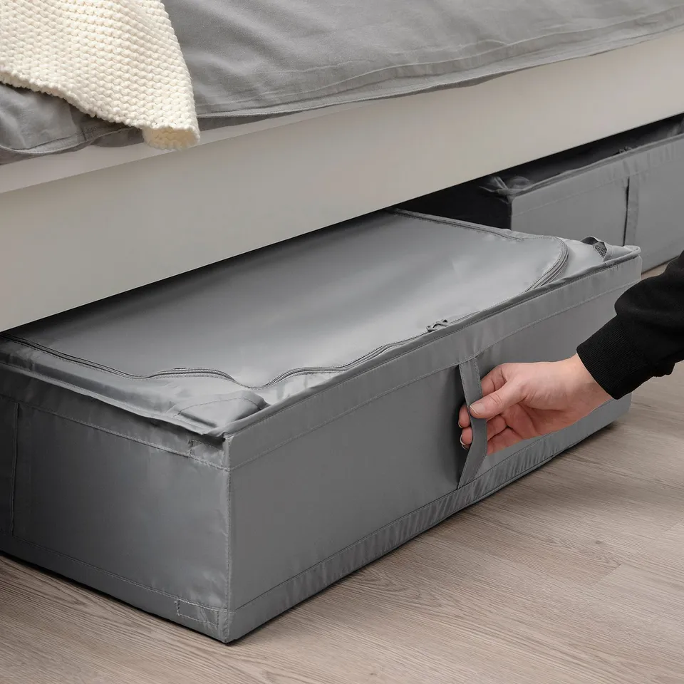 A person pulling a grey SKUBB storage case from under a bed, showcasing its use for storing clothes or bedding.