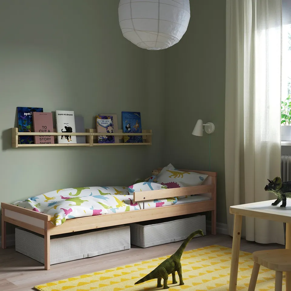 A childs room with a SNIGLAR light wood bed, colourful dinosaur bedding, and a low wooden desk. A green rug and a wall shelf with books.
