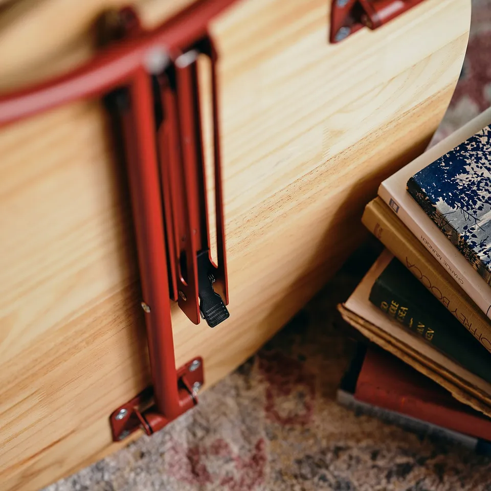 Foldable side table with vibrant red metal frame and wooden top, leaning against a stack of books.
