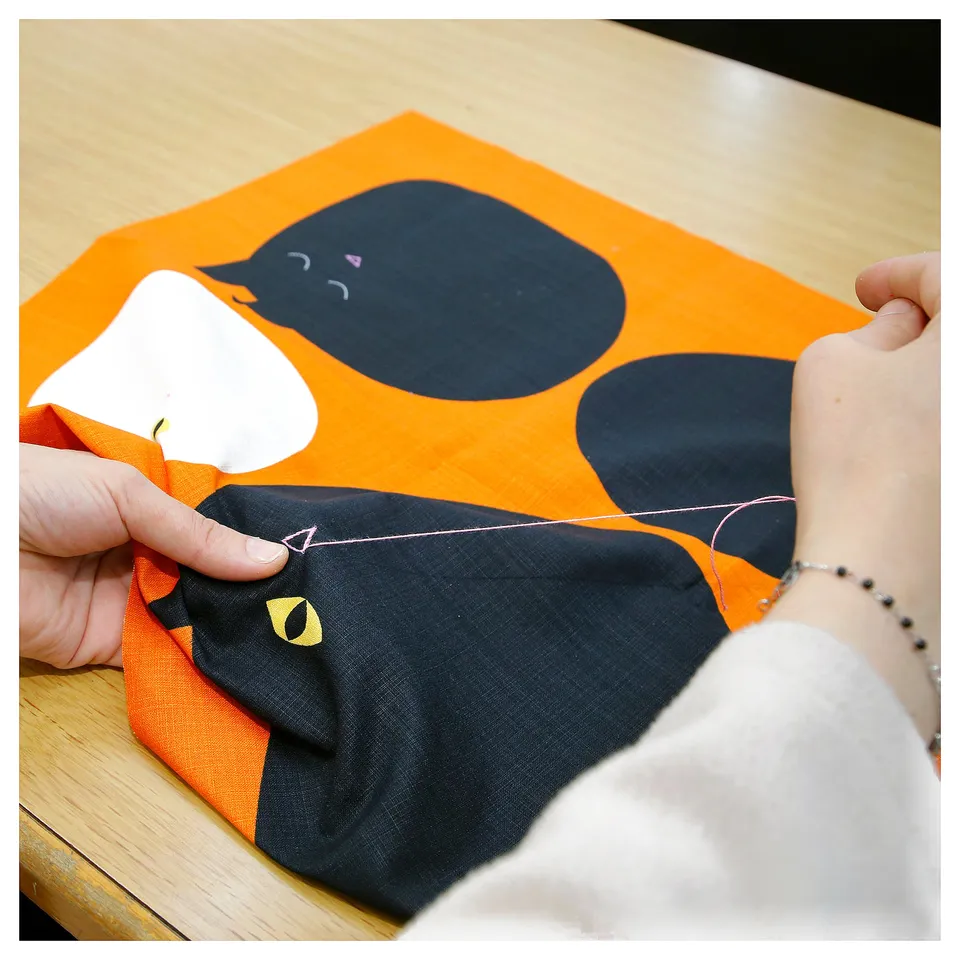 Person sewing black cat-shaped cushion on orange fabric.