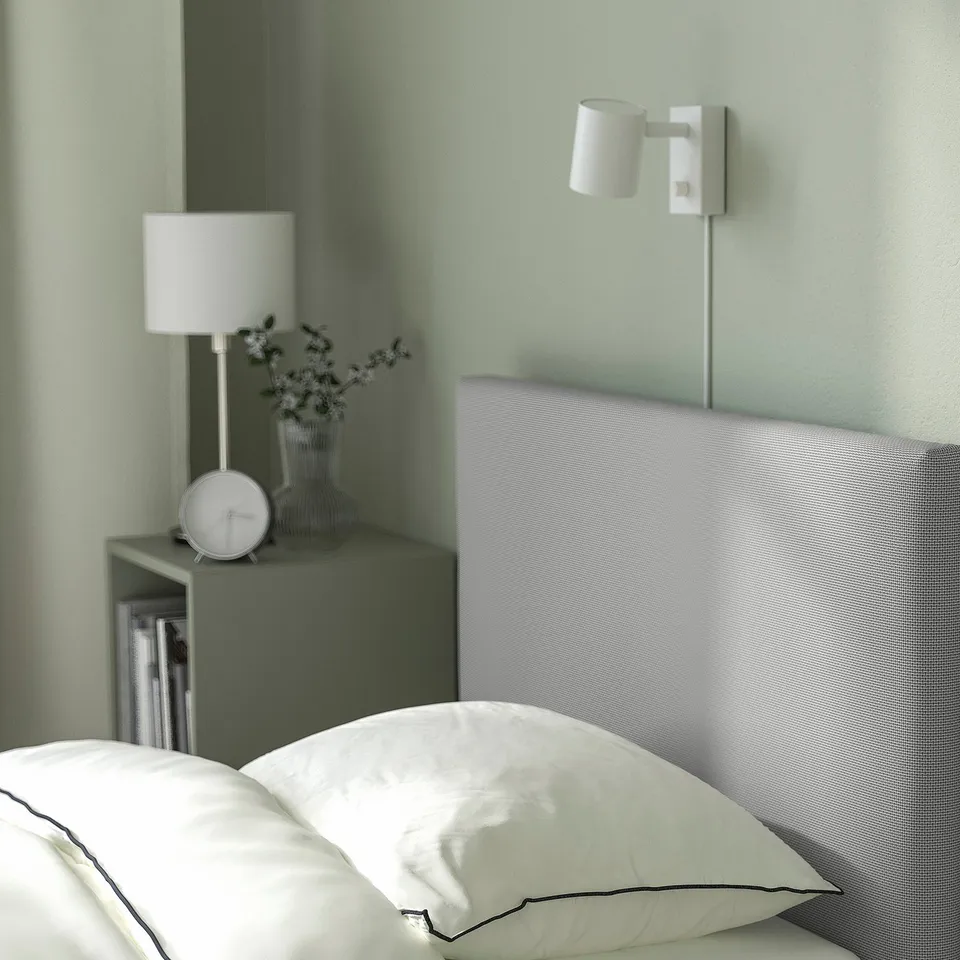 Modern bedroom with upholstered bed, white pillows, bedside lamp, vase, clock, and wall light.