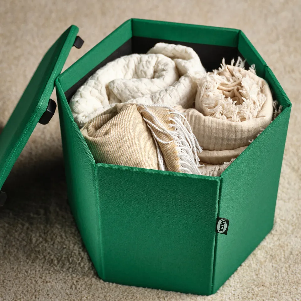 Green storage box holding folded blankets. Its part of the KJUGE multi-functional furniture series.