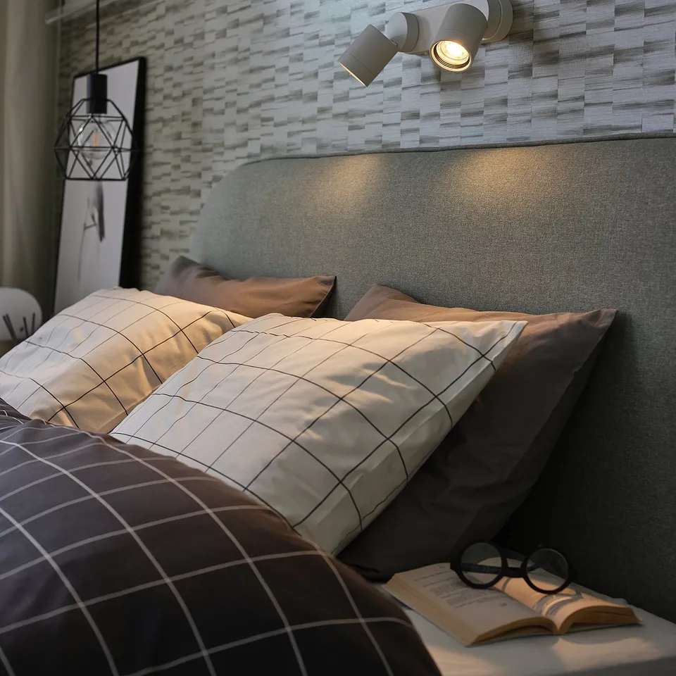 Modern bedroom with grey NYMÅNE wall lamps illuminating a bed featuring a grey headboard and patterned pillows.
