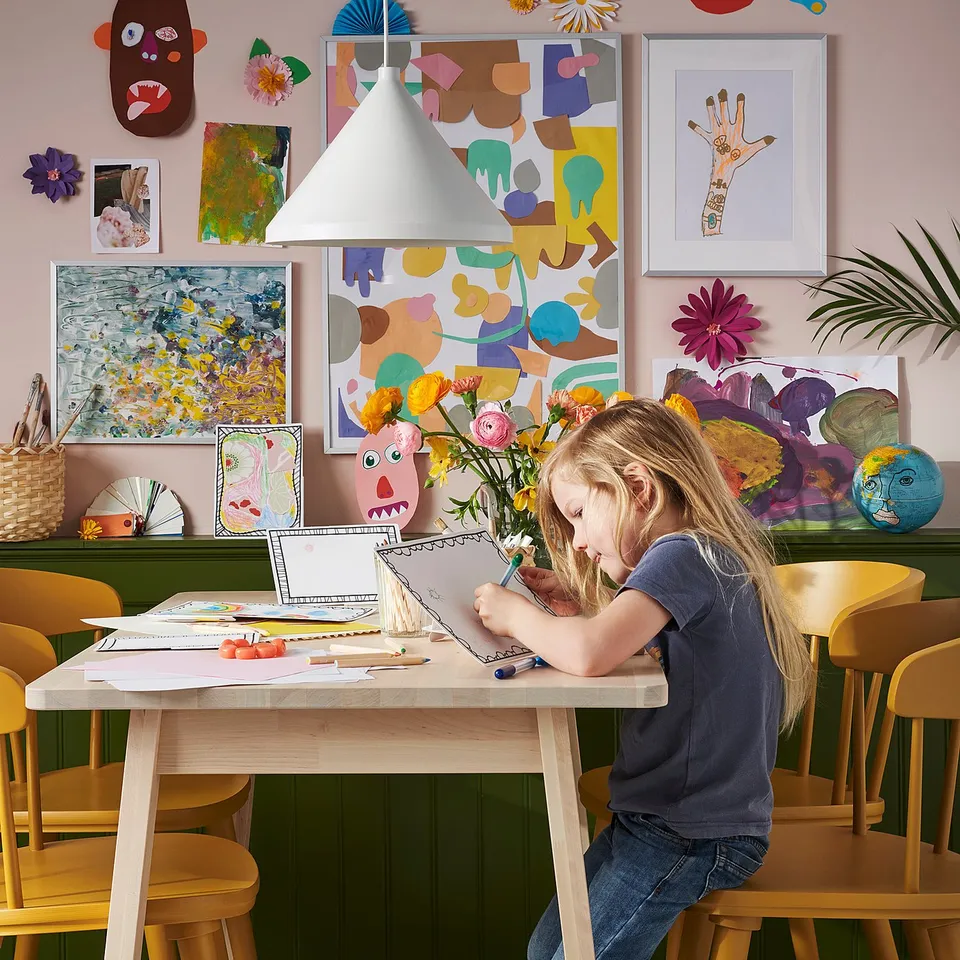 Vibrant art-filled room with child seated at natural wood NORRÅKER table, drawing. Surrounded by colourful art, masks, and hanging lights.