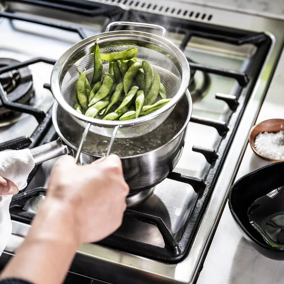 Stainless steel strainer with green beans boiling on stove.