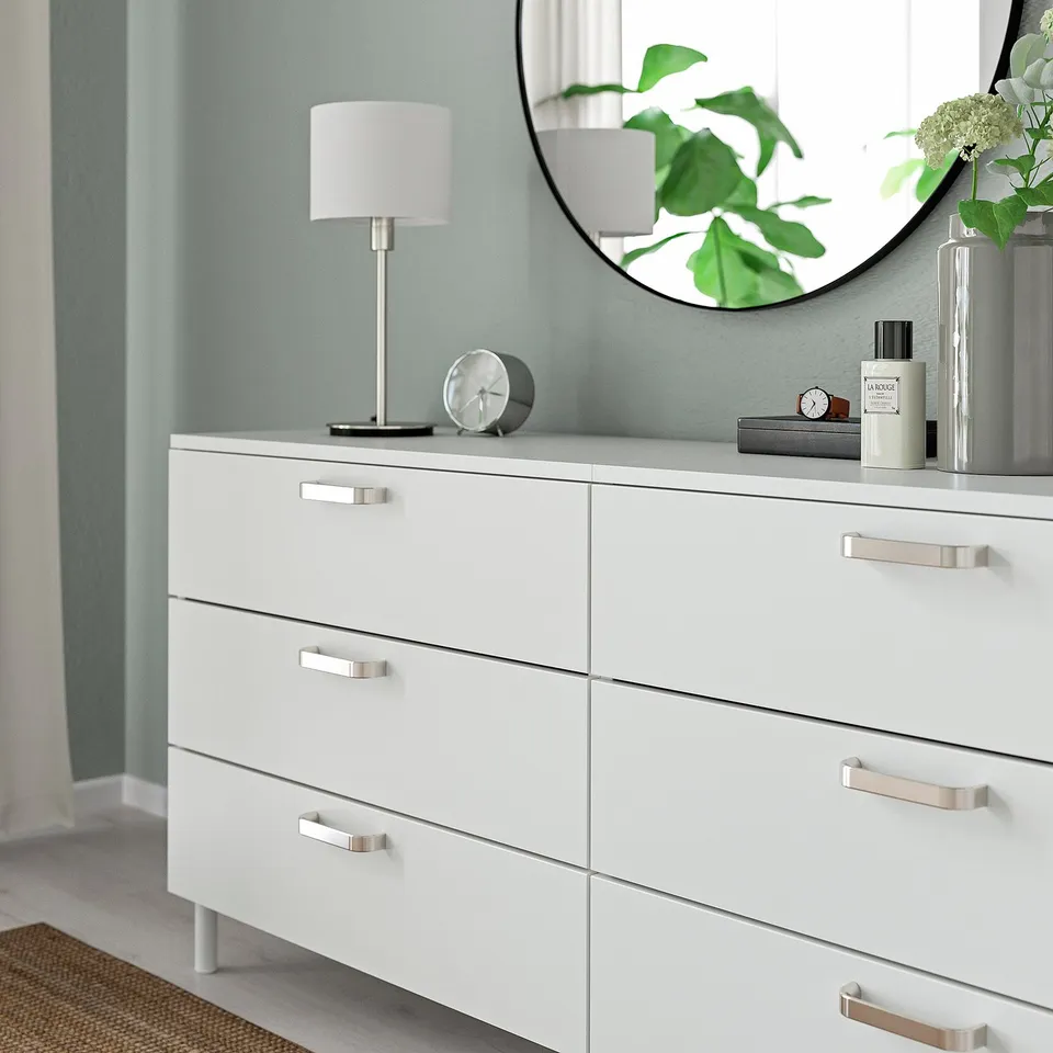 White dresser, six silver-handled drawers, lamp, mirror, and plants.