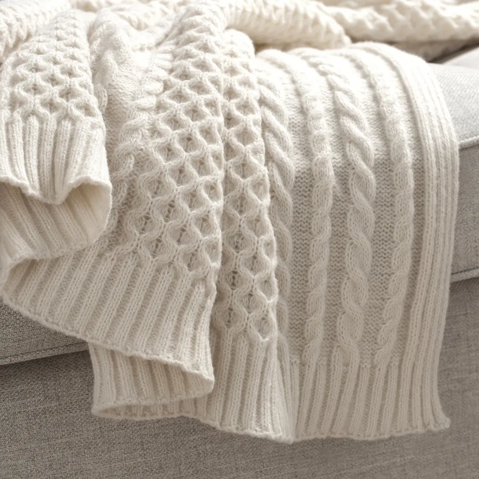 White knitted throw blanket with intricate cable patterns, draped over a grey couch.