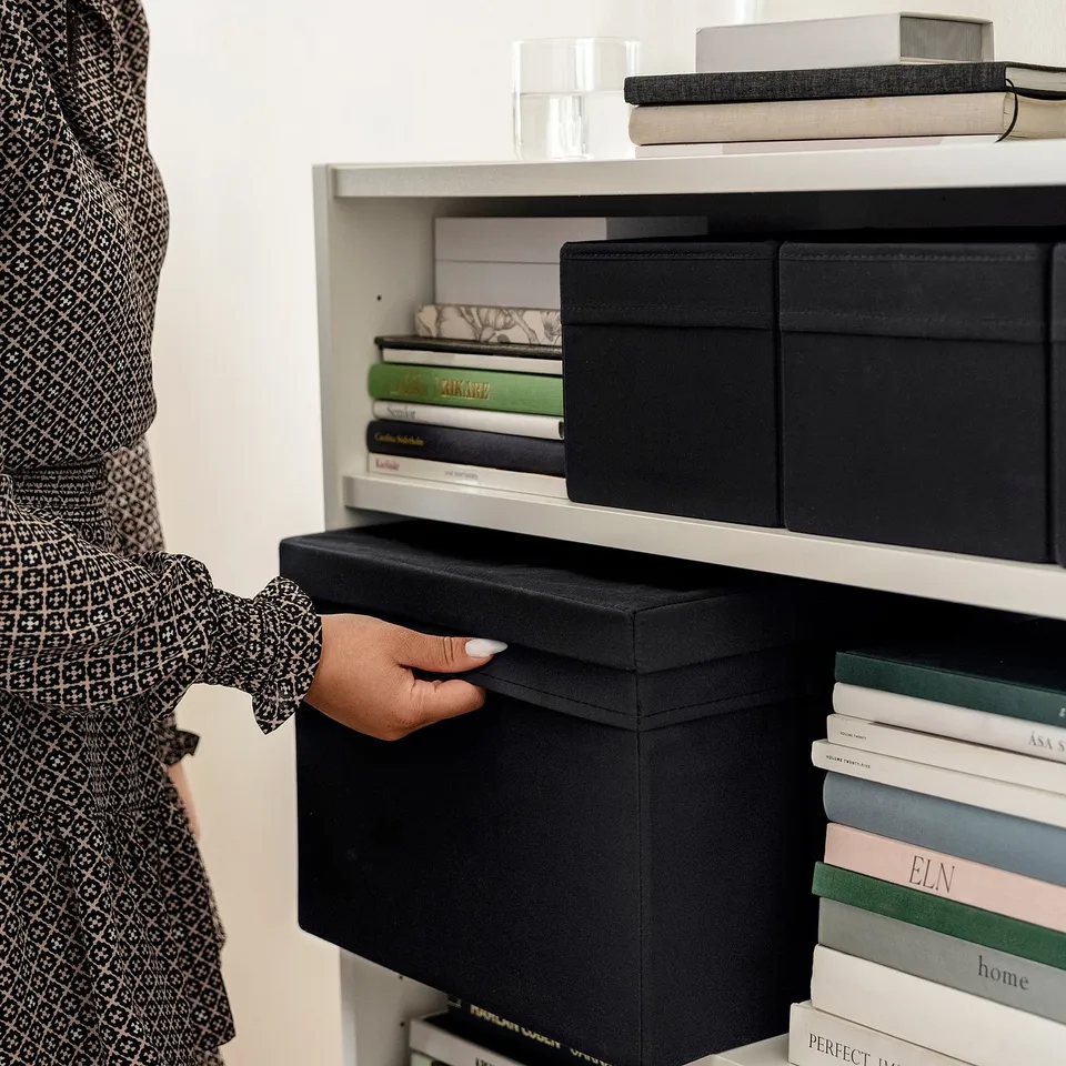 Person organising black fabric-covered boxes on a white shelf. The boxes fit perfectly and are easy to fold and unfold.