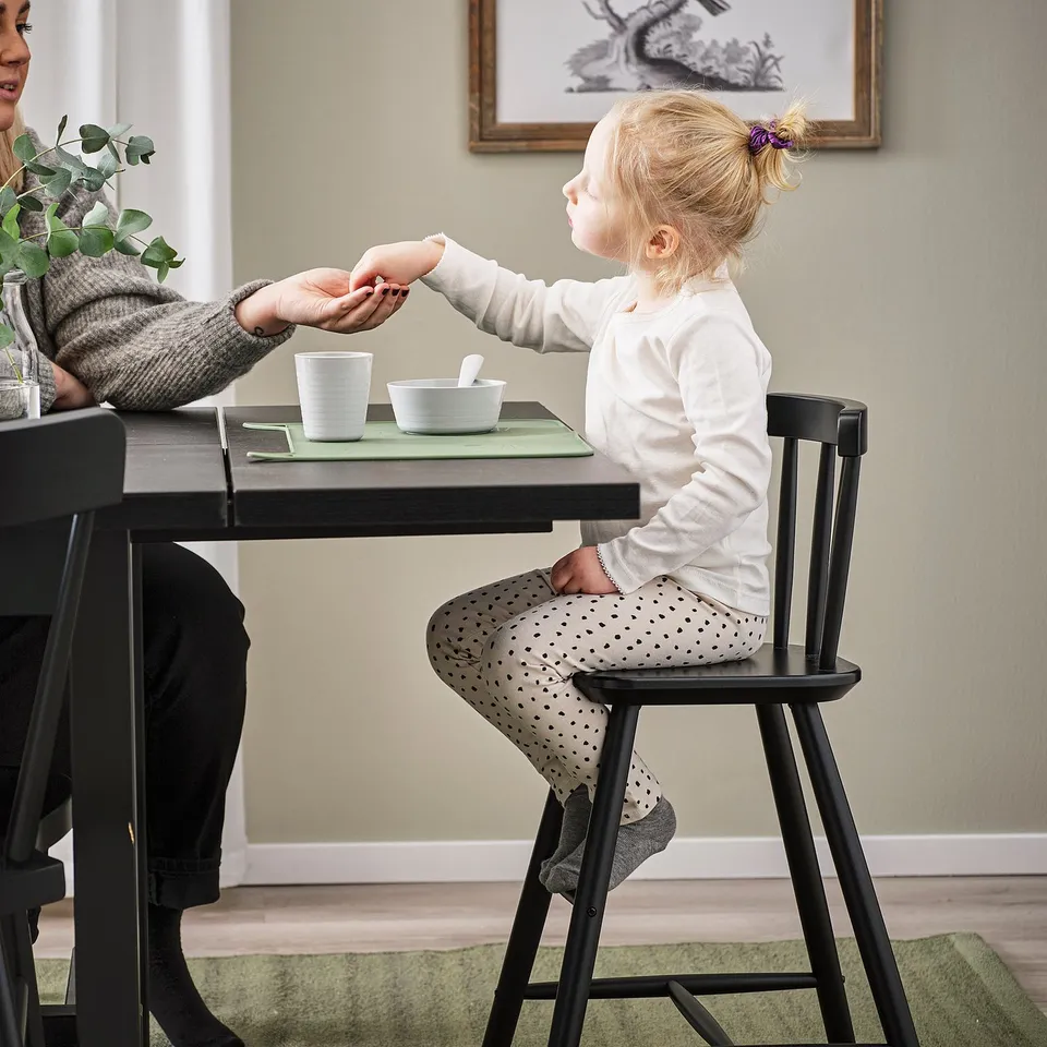 Young child sitting on a junior chair, eating from a bowl with a spoon. The chair provides proper height and foot support for dining.