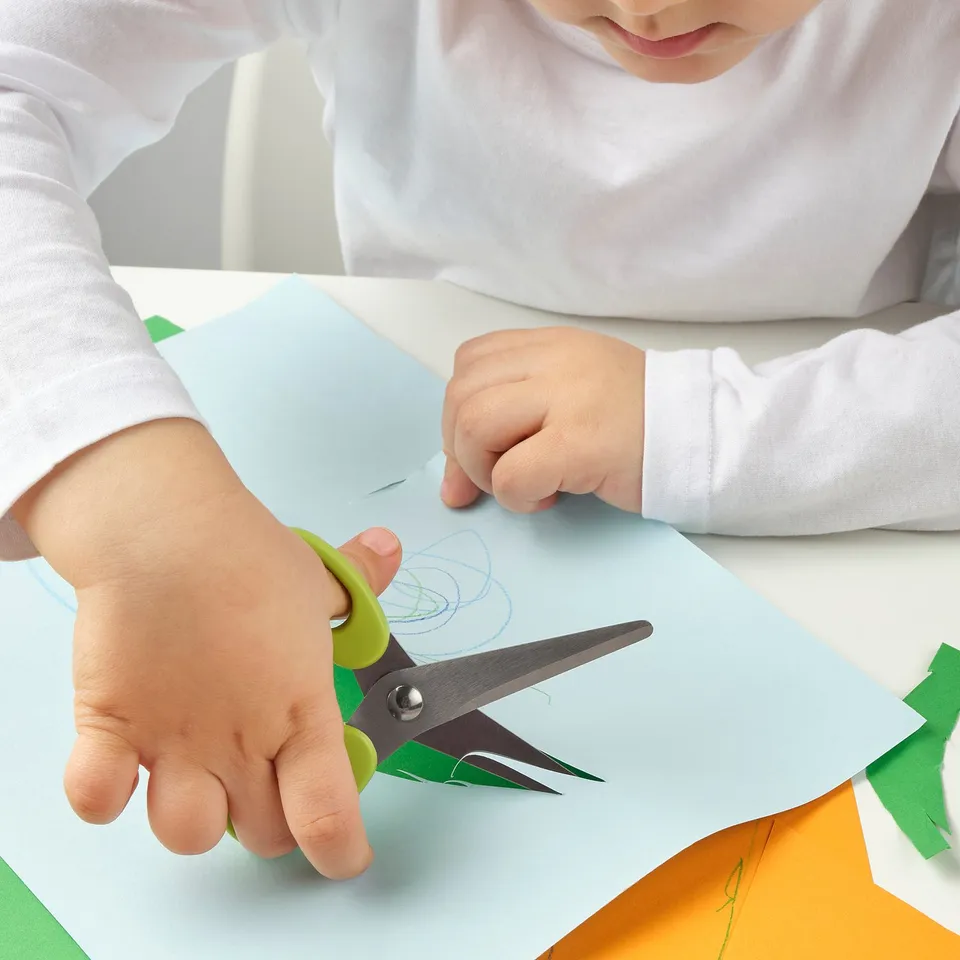 Child safely cuts blue paper with green-handled scissors.