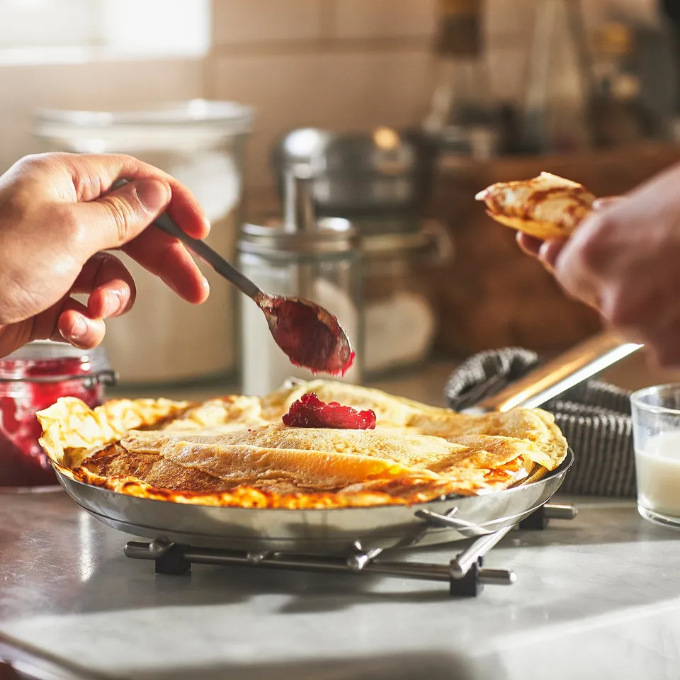 A person flips a pancake in a grey pan on a stove, with red jam being spooned onto another pancake.