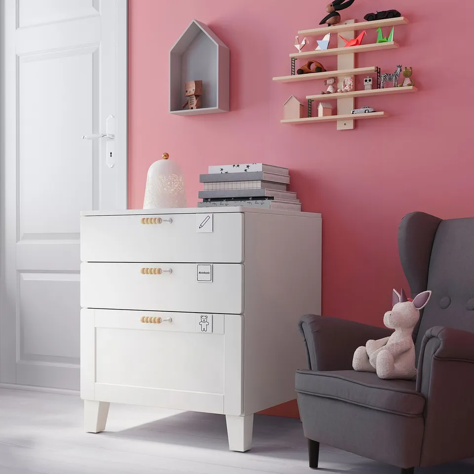 Small childrens room with pink walls, white SMÅSTAD chest of drawers, grey chair, stuffed unicorn, books, and toys on shelves.