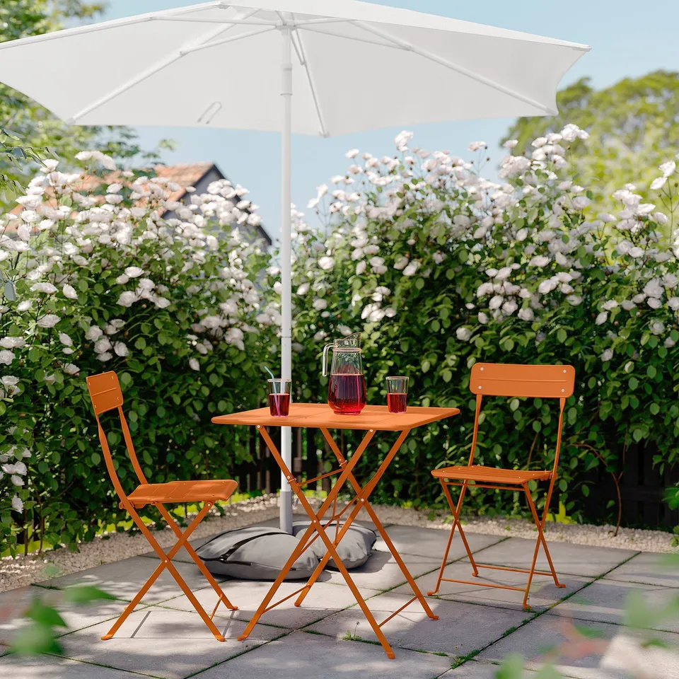 Small outdoor setting with orange foldable chairs and table under a white umbrella. Surrounded by white flowers.