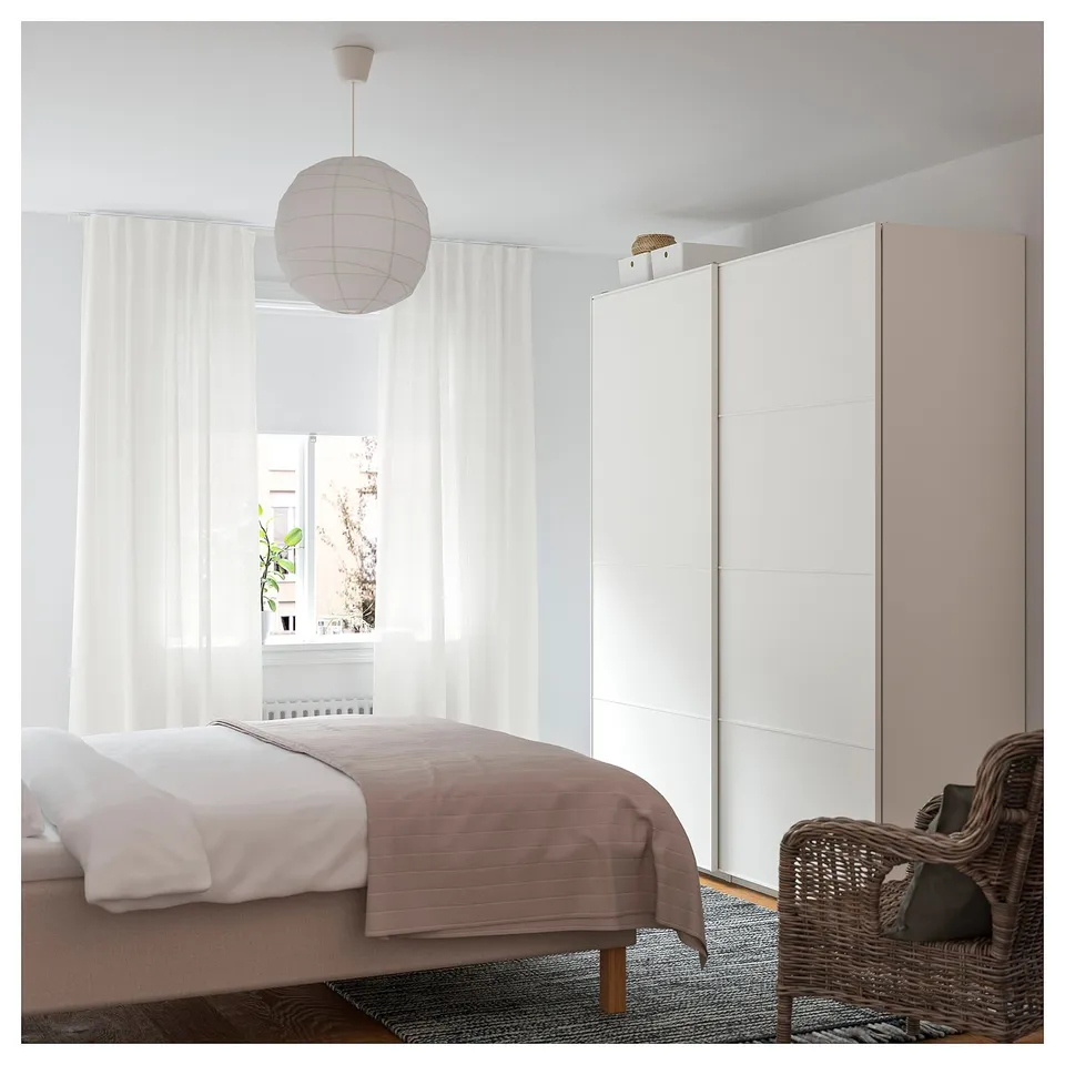 Bright, minimalist bedroom with white walls, a grey bedspread, hanging white pendant light, wicker chair, and large window with curtains.