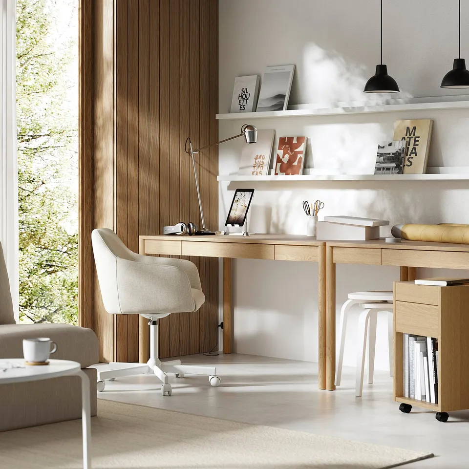Modern home office with light oak TONSTAD desk, white chair, shelves with books, and grey couch. Desk is neat with a computer, lamp, and stool.