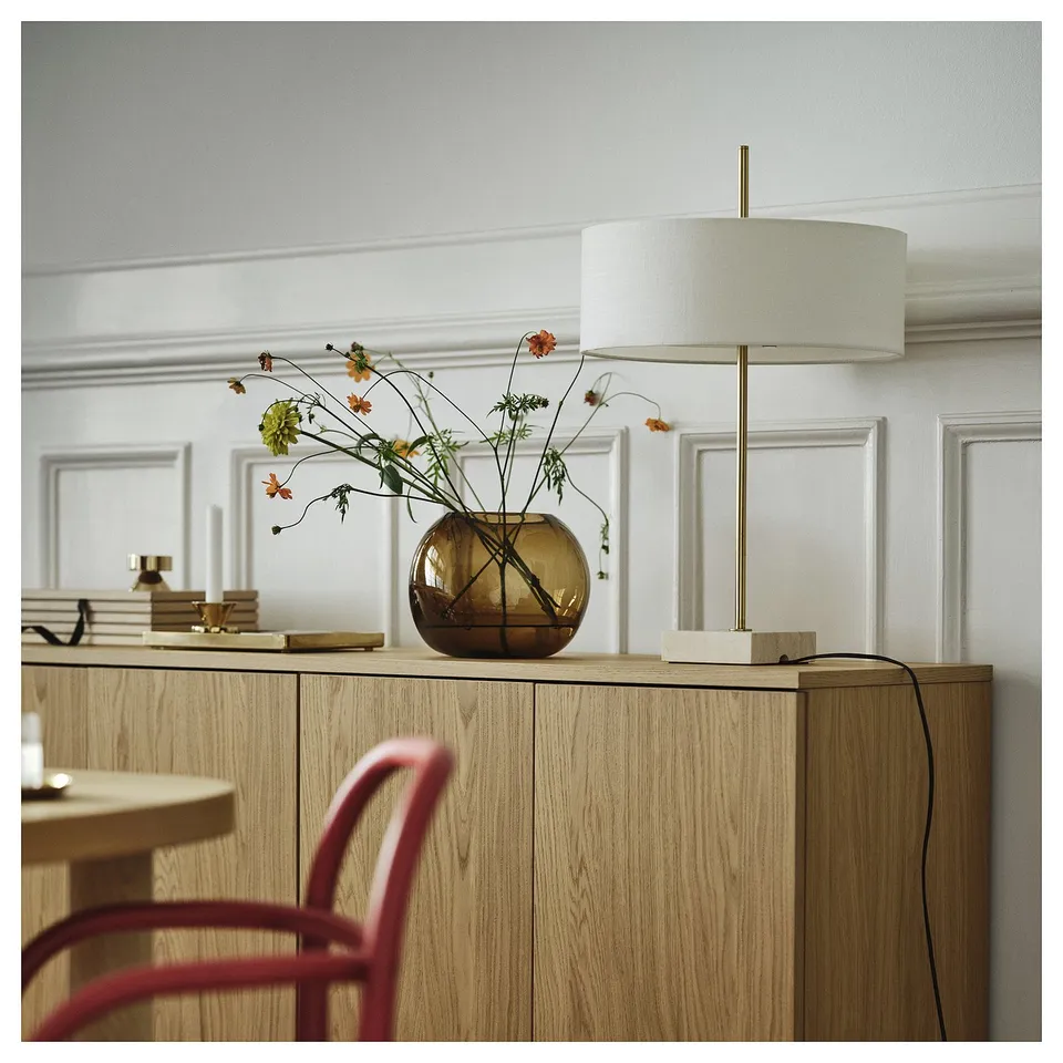 Modern lamp with a brass-colored base and white shade sits on a wooden sideboard next to a flower vase. The lamps size is similar to the vase.